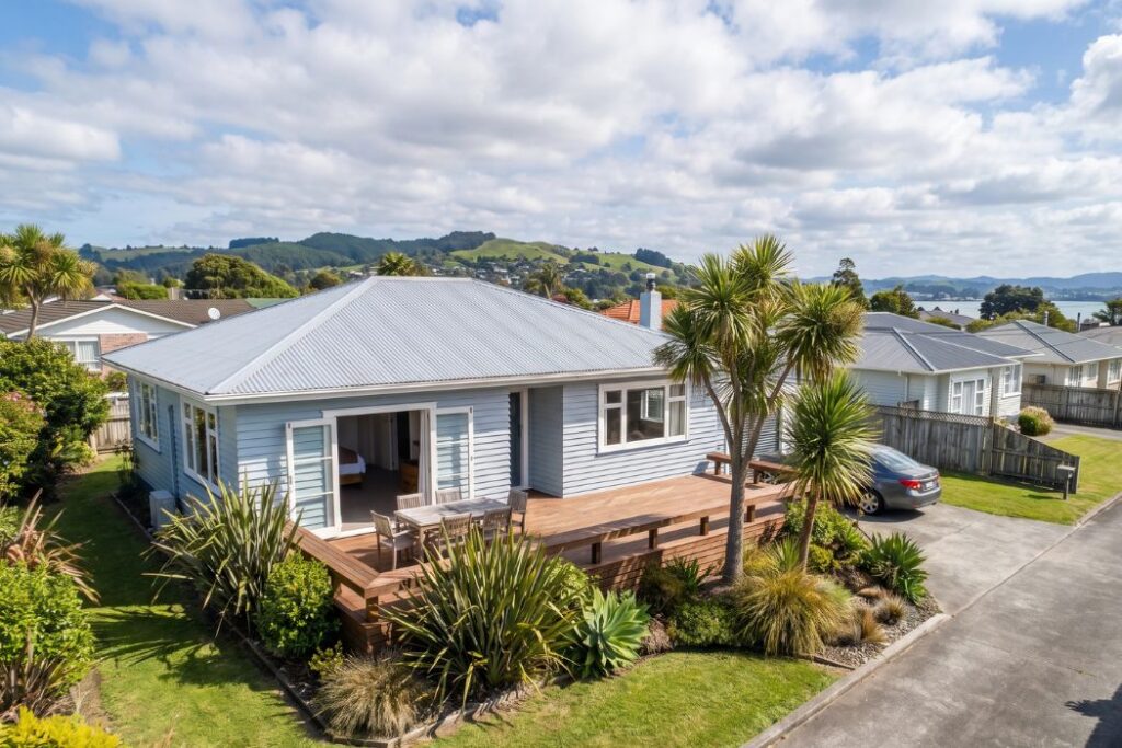 A blue weatherboard New Zealand home with a deck and cabbage trees, overlooking the harbour in the Bay of Plenty